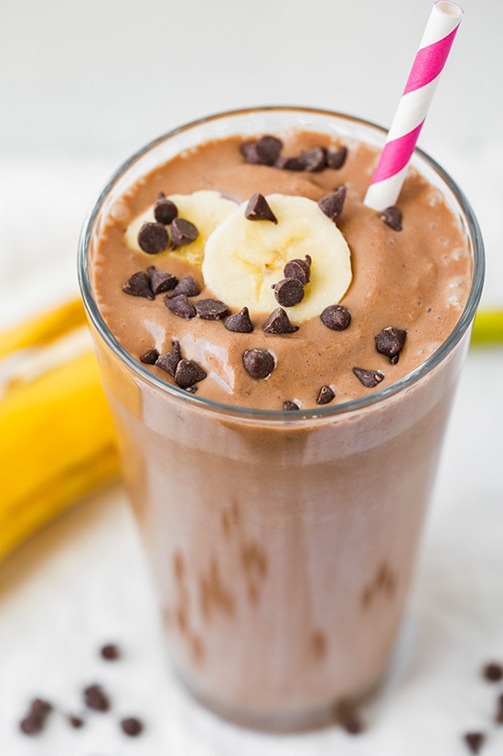 A Banana Chocolate smoothie in a tall glass topped with chocolate chips, banana slices, and a white/pink swirl straw
